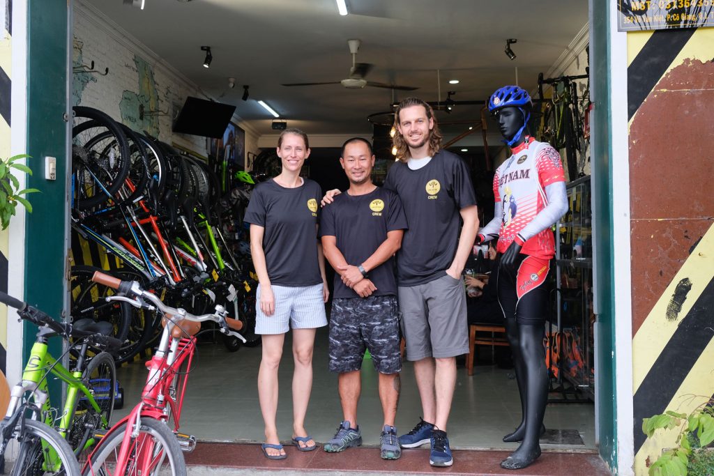 Leo und Sebastian stehen mit einem Mann vor einem Fahrradgeschäft in Ho-Chi-Minh-Stadt.